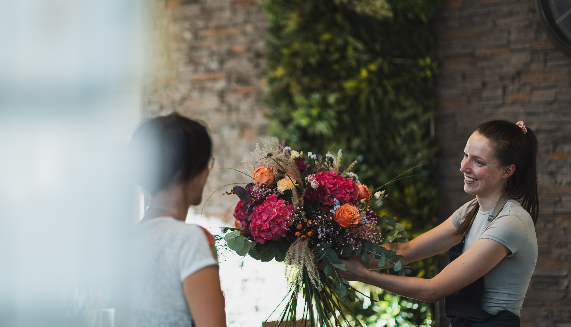 Eine lächelnde Floristin überreicht einer Kundin in einem Geschäft mit grüner Pflanzenwand einen großen, bunten Blumenstrauß. - Obojes