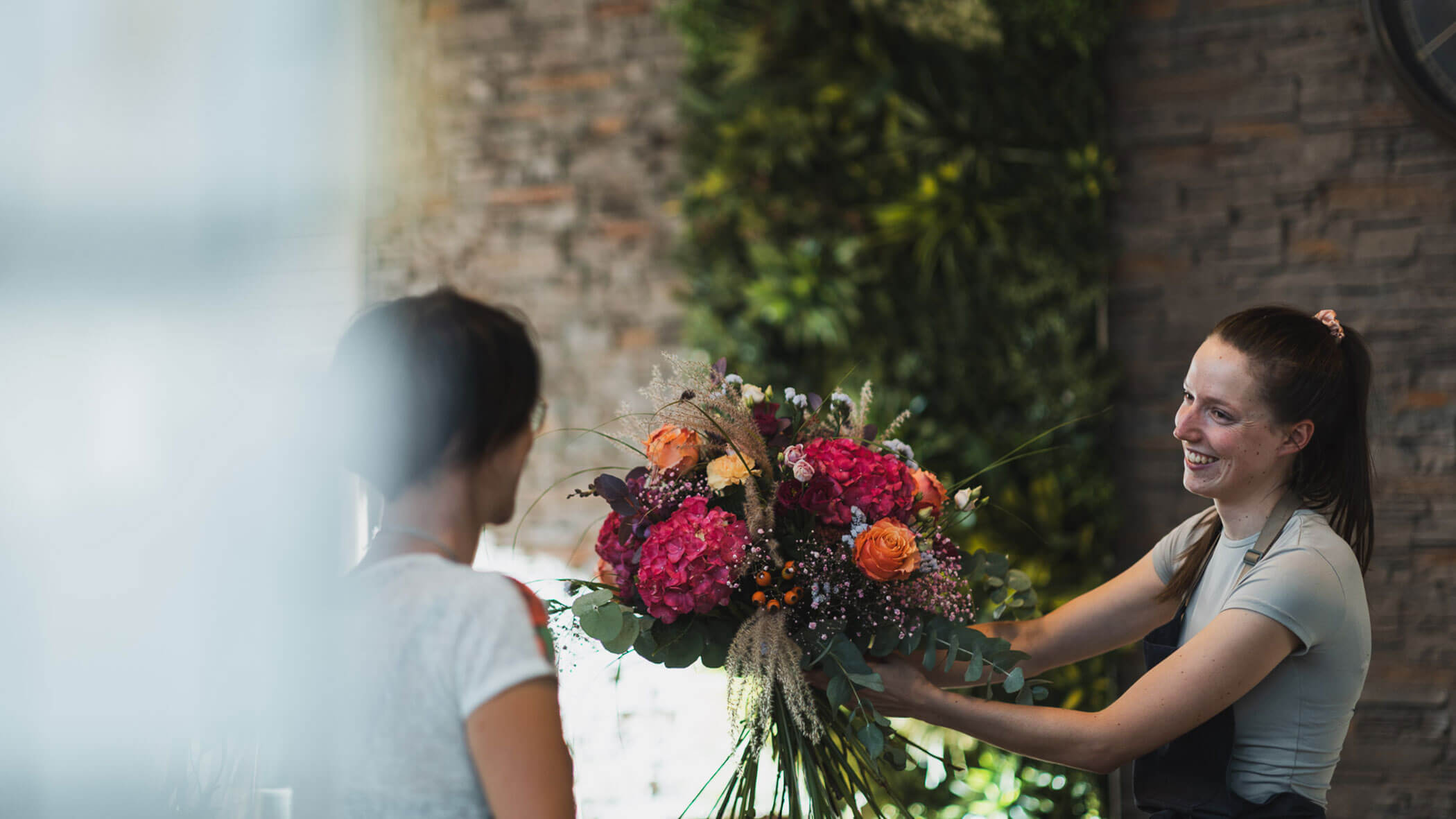 Eine lächelnde Blumenhändlerin überreicht einer Kundin einen großen, bunten Blumenstrauß in einem Geschäft mit grünen Wänden. - Obojes