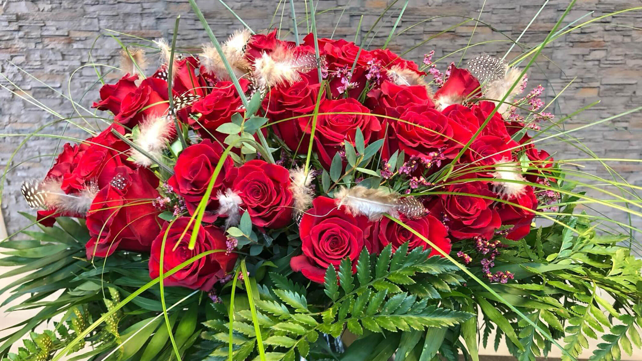 Ein üppiger Strauß roter Rosen, Grünzeug, Gras, Federn und kleine rosa Blumen vor einem Hintergrund aus Steinmauern. - Obojes