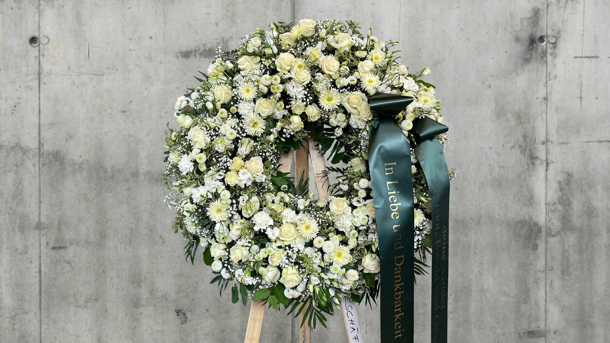 Großer weißer und cremefarbener Blumenkranz mit grünen Bändern, der an einer grauen Betonwand angebracht ist. - Obojes