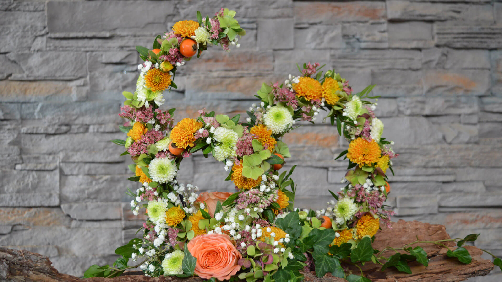 Blumengesteck in Form der Zahl 80 mit orangefarbenen, weißen und grünen Blumen vor einer Steinmauer. - Obojes
