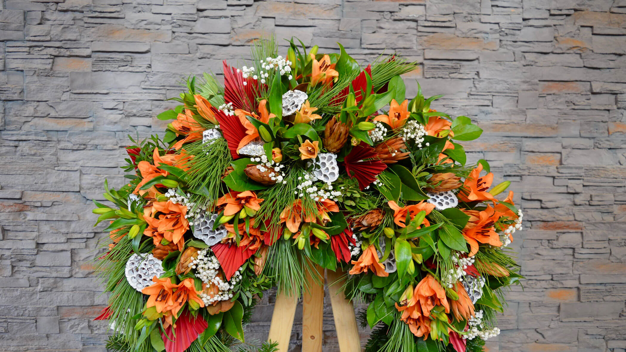 Ein Trauerkranz mit orangefarbenen Blumen, Grünzeug und weißen Akzenten vor einem Steinmauer-Hintergrund. - Obojes