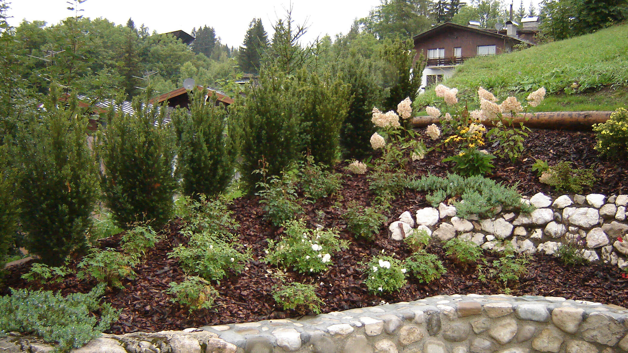 Ein Garten am Hang mit Sträuchern, Blumen, Steinmauern und Häusern im Hintergrund an einem bewölkten Tag. - Obojes