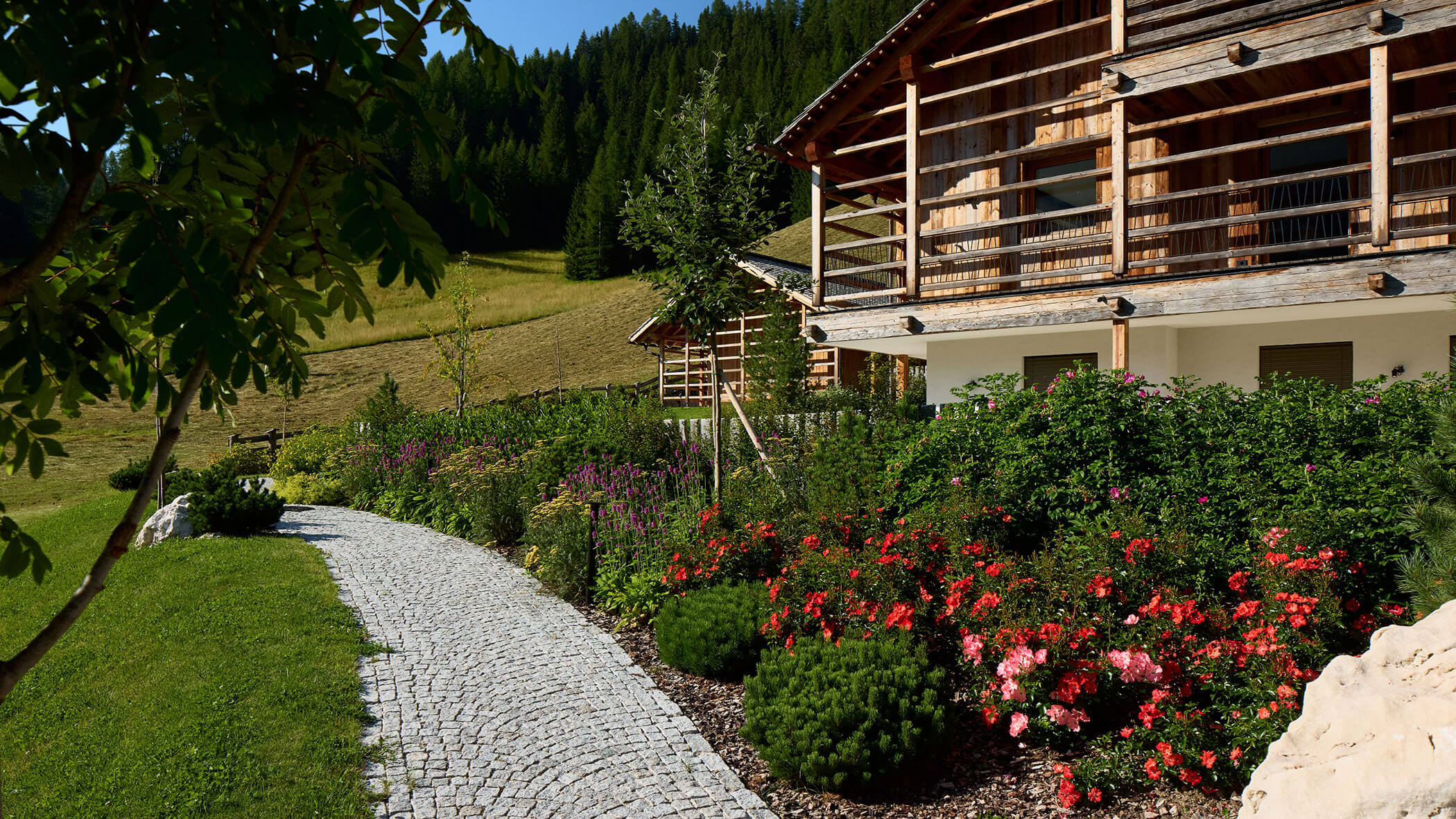 Ein Steinweg schlängelt sich vorbei an blühenden Blumen und einem Holzhaus in einer sonnigen, grünen Berglandschaft. - Obojes