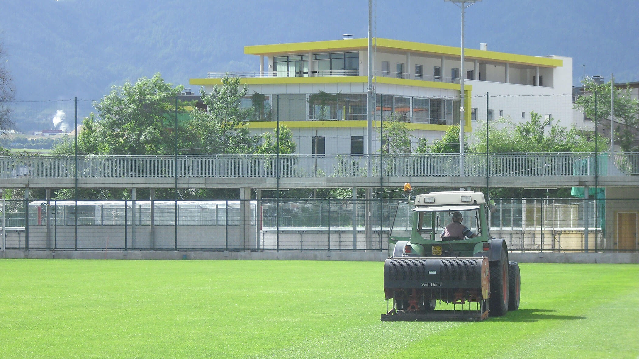 Eine Person fährt einen Rasenbelüfter auf einem hellgrünen Sportplatz in der Nähe eines modernen Gebäudes und von Bergen. - Obojes