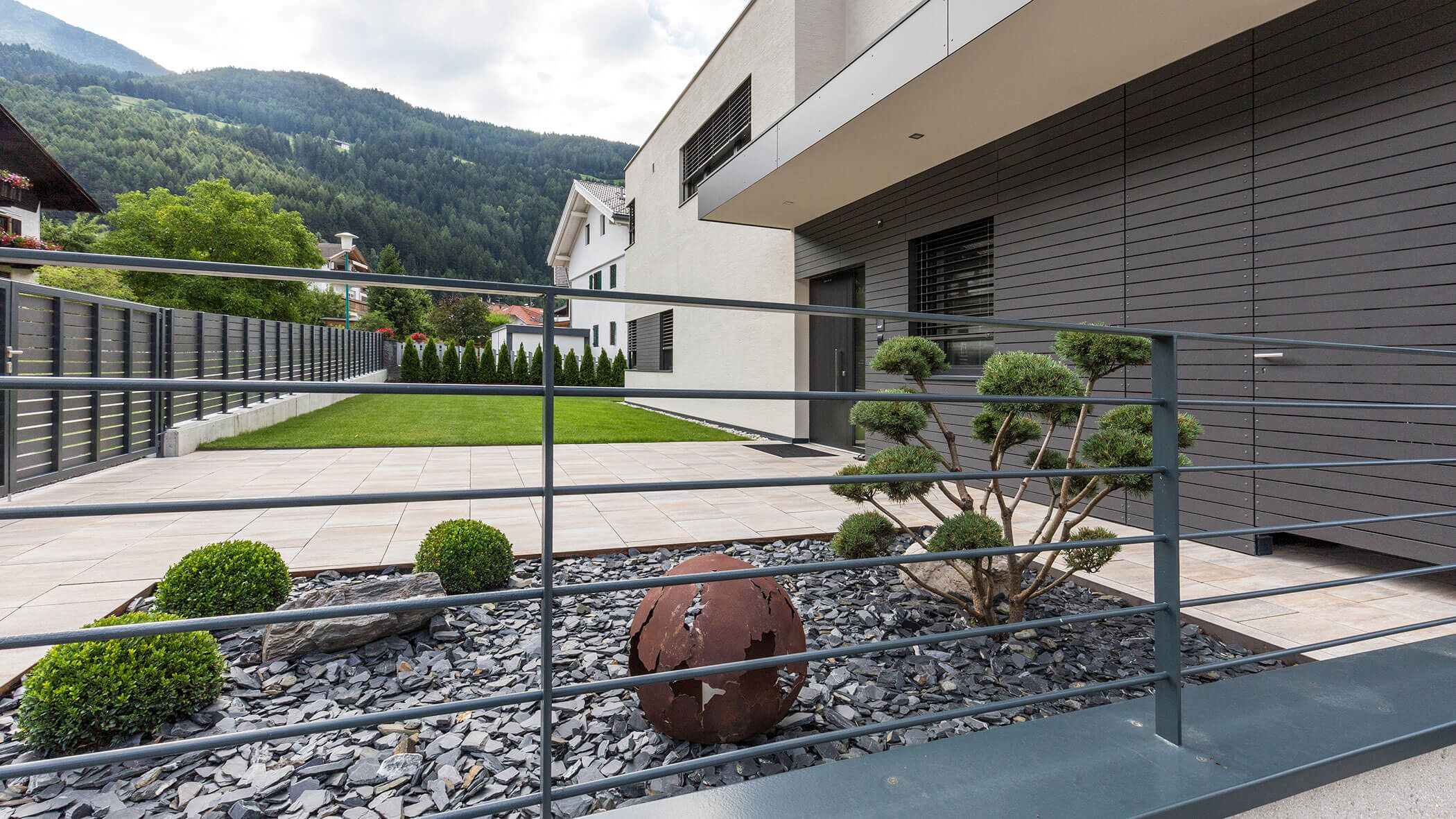 Moderne Hausfassade mit gepflegtem Rasen, dekorativen Steinen, Sträuchern und einer Metallkugel-Skulptur vor dem Haus. - Obojes