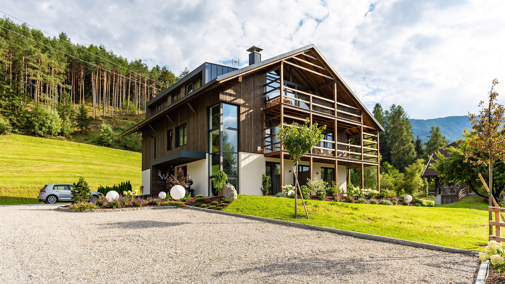 Modernes Holzhaus mit großen Fenstern und Balkonen, umgeben von Grün und Bergen unter einem teilweise bewölkten Himmel. - Obojes
