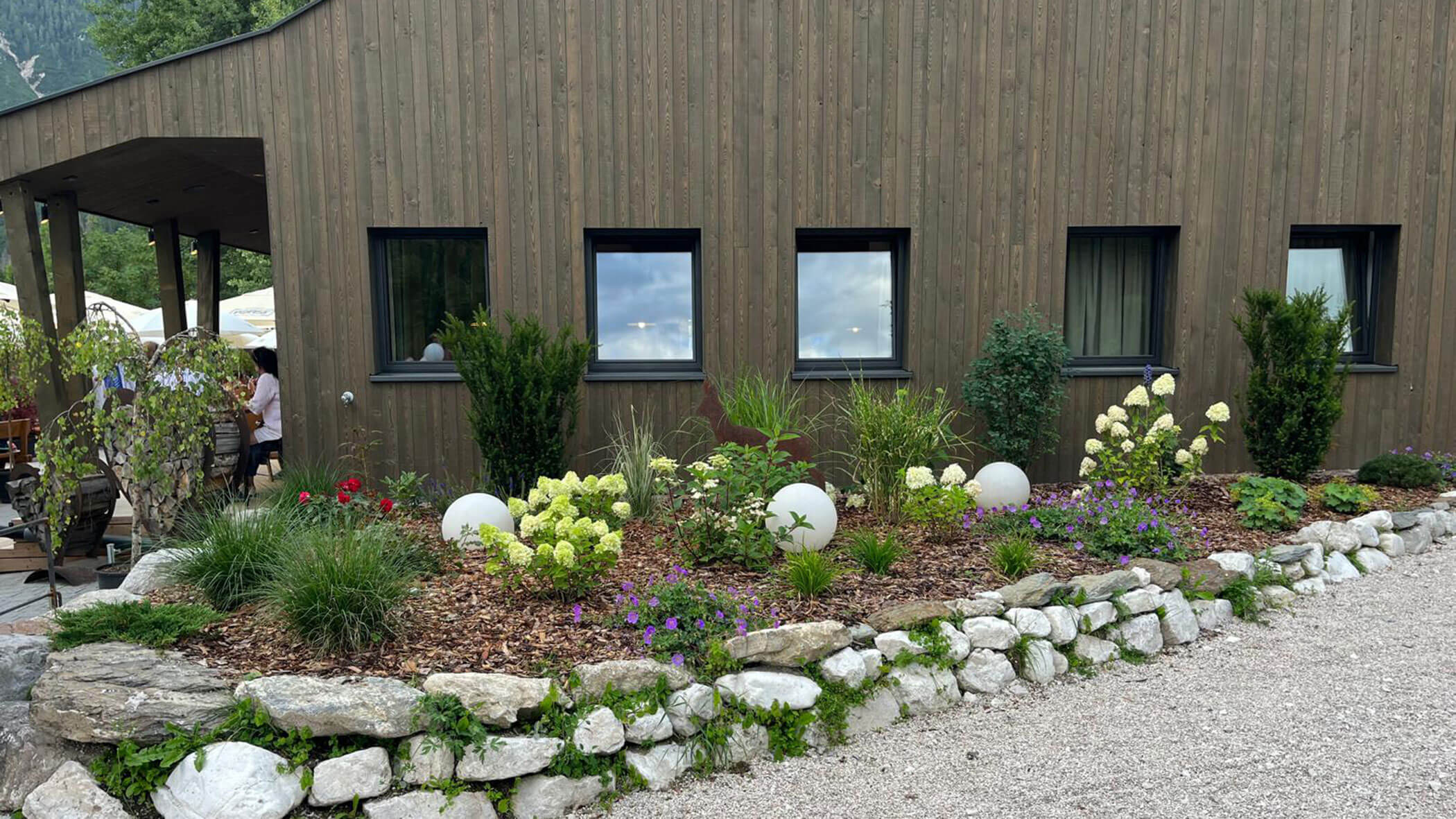 Ein modernes Holzgebäude mit einem angelegten Garten mit Blumen, Sträuchern, Steinen und weißen kugelförmigen Ornamenten. - Obojes
