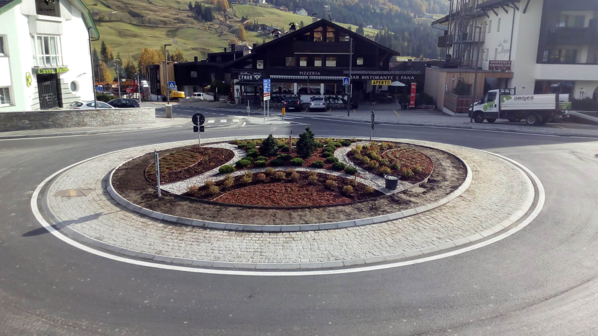 Ein Kreisverkehr mit Pflanzen und Wegen, umgeben von Gebäuden in einer bergigen Gegend. - Obojes