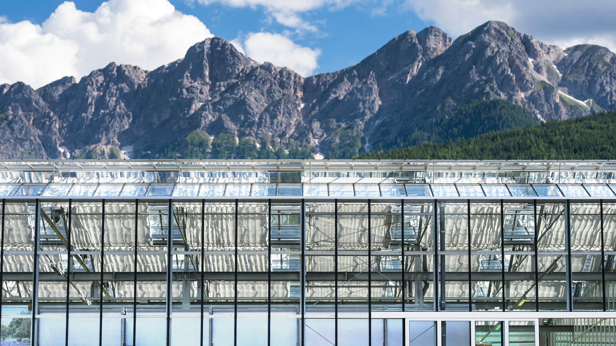 Gläsernes Gewächshaus vor hohen, felsigen Bergen unter einem teilweise bewölkten blauen Himmel. - Obojes