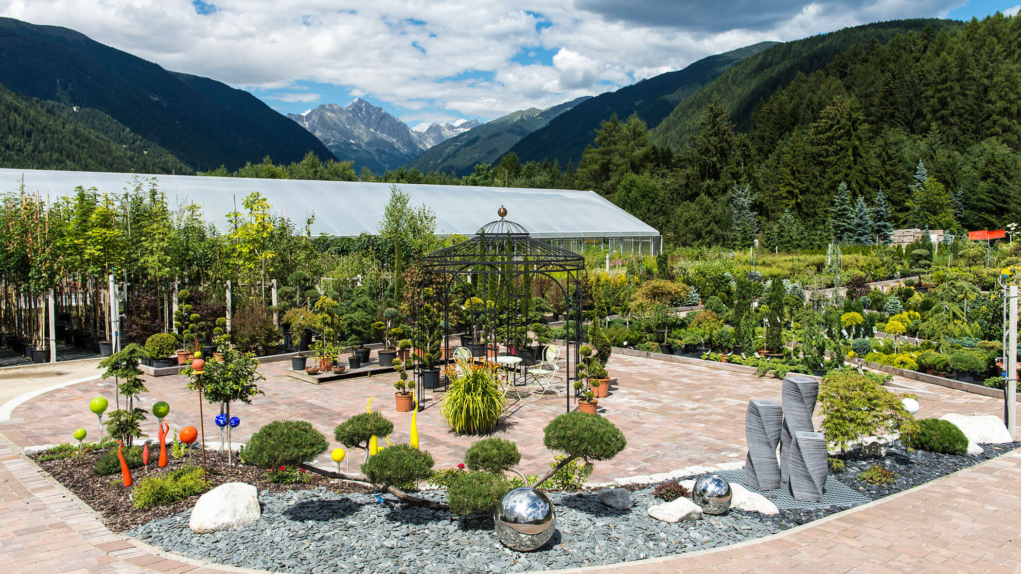 Garten mit Pflanzen, Skulpturen und einem Pavillon vor der Kulisse von Bergen und einem teilweise bewölkten Himmel. - Obojes