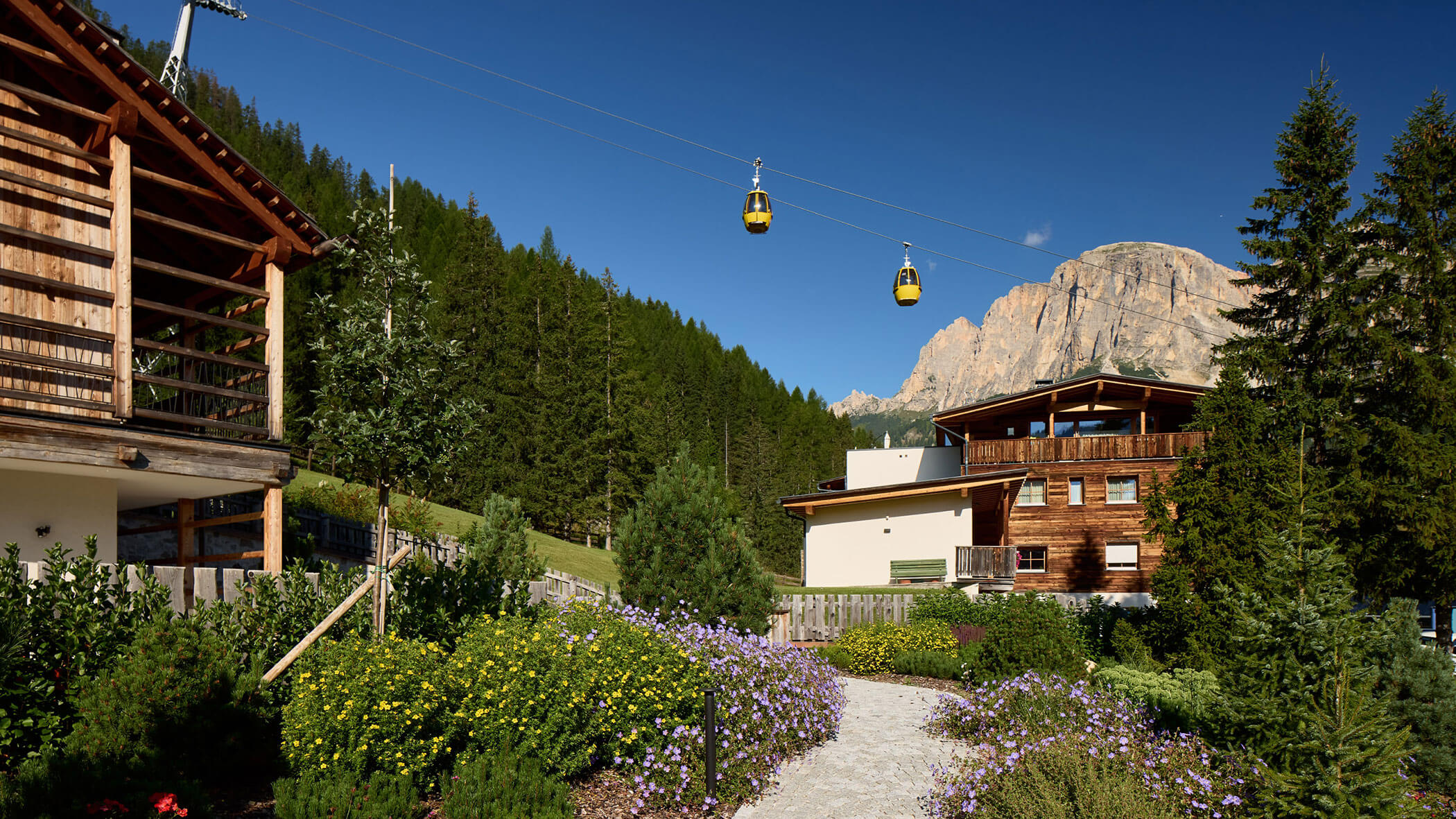 Holzhütten und Gärten mit Seilbahnen darüber, Wald und ein felsiger Berg im Hintergrund unter blauem Himmel. - Obojes