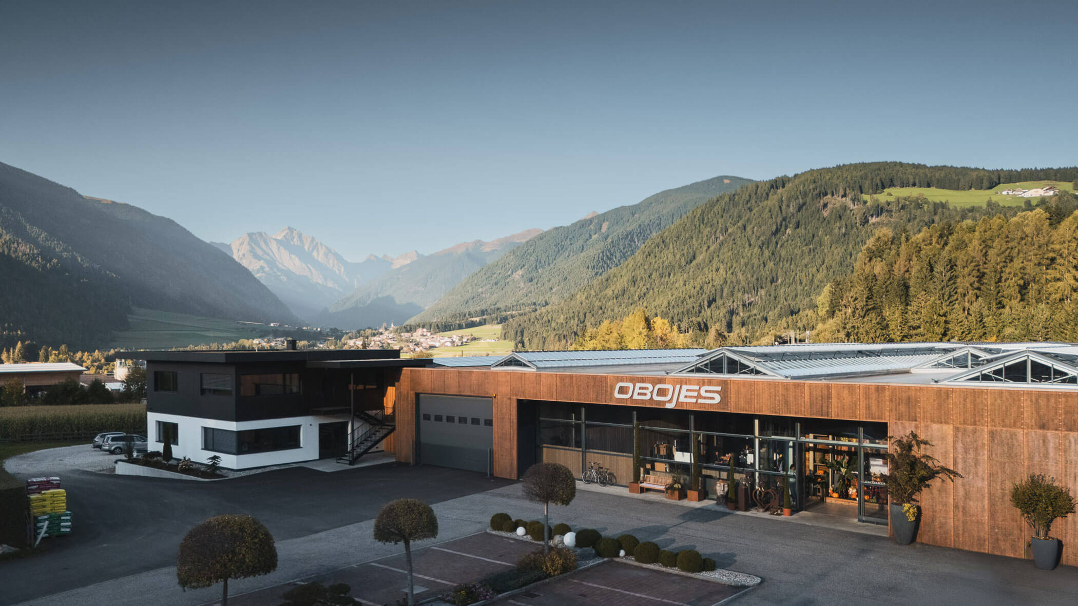 Modernes Gebäude mit der Aufschrift OBOJES mit Bergen und Bäumen im Hintergrund unter einem klaren Himmel. - Obojes