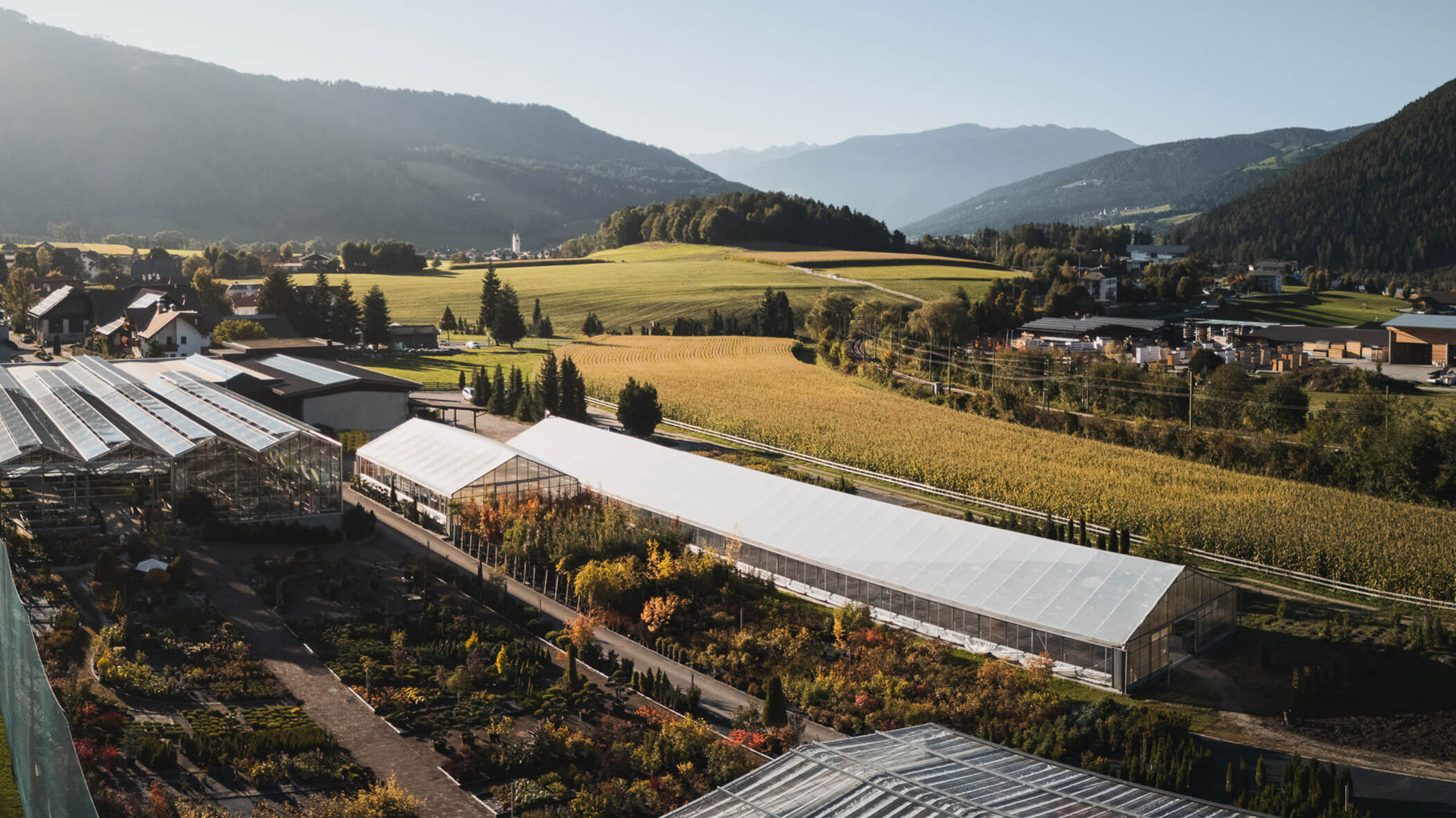 Luftaufnahme von Gewächshäusern und Feldern, umgeben von Bergen und einem kleinen Dorf unter einem klaren Himmel. - Obojes